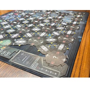 May include: A board game with a dark blue and grey colour scheme. The game board is covered with hexagonal tiles and includes informational cards with text. The game appears to be a strategy or adventure game.