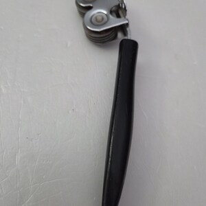 May include: A vintage can opener with a black handle and a silver-colored, multi-roller mechanism. The can opener is designed to cut the top of a can. The handle is smooth and ergonomic.