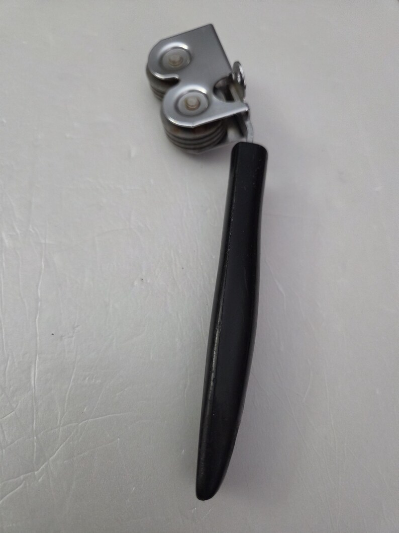 May include: A vintage can opener with a black handle and a silver-colored, multi-roller mechanism. The can opener is designed to cut the top of a can. The handle is smooth and ergonomic.