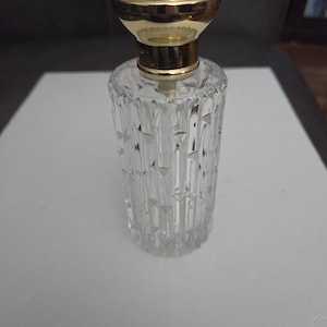 May include: A clear glass perfume bottle with a gold-colored cap. The bottle has a textured, vertical design. The cap is round with a decorative pattern. The bottle is empty and sits on a white surface.
