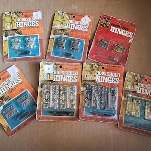 Peut inclure: Une collection de charnières Homecraft et décoratives vintage de différents styles. Les charnières sont emballées sur un support en carton orange et rouge avec le nom du produit et les dimensions. Certaines charnières sont argentées, dorées et bronze.