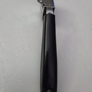 May include: A metal can opener with a black handle. The can opener has a silver-colored, rectangular head with two small, circular holes on the top. The handle is black, with a hole at the bottom for hanging.