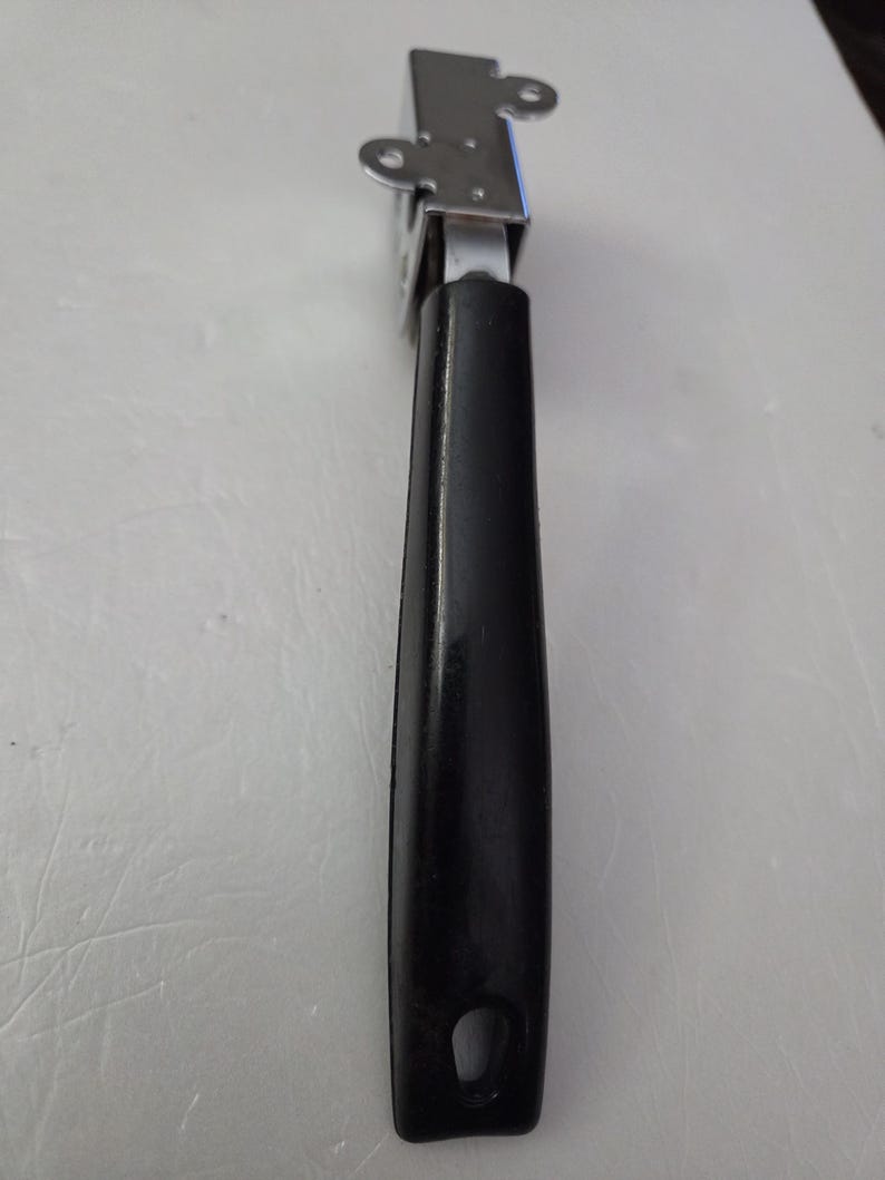 May include: A metal can opener with a black handle. The can opener has a silver-colored, rectangular head with two small, circular holes on the top. The handle is black, with a hole at the bottom for hanging.