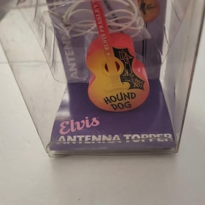 May include: An Elvis antenna topper in a clear plastic package. The guitar-shaped topper is orange and yellow with the words "HOUND DOG" and "ELVIS PRESLEY" on it. The package also says "ANTENNA TOPPER".