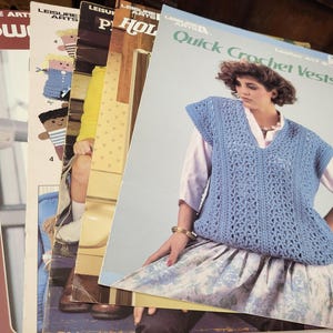 May include: A stack of vintage crochet pattern books. The top book features a woman wearing a blue crochet vest. Other books show illustrations of crochet projects. The books are from the Leisure Arts series.