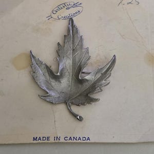 May include: A silver-toned metal maple leaf brooch attached to a beige display card. The card features the text Artistic Creations at the top, the handwritten number 1.25 in the corner, and the text MADE IN CANADA at the bottom.