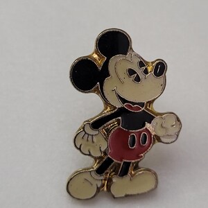 May include: A gold-toned enamel pin featuring Mickey Mouse. The pin depicts Mickey in his classic pose, with black ears, a cream-colored face and hands, a red shirt, and black shorts. The pin has a gold border.