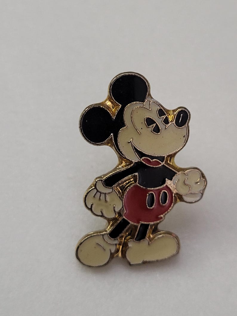 May include: A gold-toned enamel pin featuring Mickey Mouse. The pin depicts Mickey in his classic pose, with black ears, a cream-colored face and hands, a red shirt, and black shorts. The pin has a gold border.