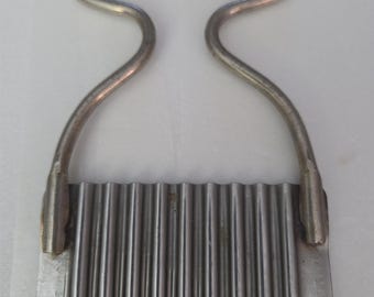 Op de afbeelding: Een zilverkleurige metalen rimpelsnijder met een golvend, geribbeld mes bevestigd aan een afgerond, boogvormig handvat. Het gereedschap is gemaakt van roestvrij staal en heeft een getextureerde snijrand voor het snijden van groenten.