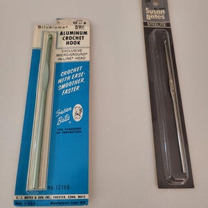 May include: Two crochet hooks packaged in plastic. The hook on the left is a silver aluminum crochet hook size 6, made by Susan Bates. The hook on the right is a silver steel crochet hook size 7, also made by Susan Bates.