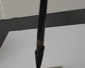 May include: A black pen in a marble pen holder. The pen is dark with a gold-colored band and a brass-colored base. The pen holder is rectangular with a marbled, off-white and gray pattern.