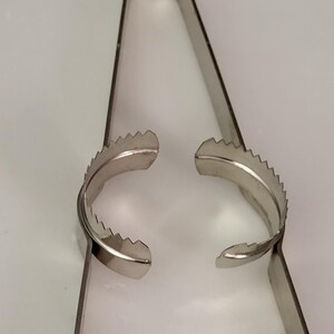 Puede incluir: Un par de pinzas de metal plateado con bordes dentados en los extremos. Las pinzas est&aacute;n abiertas y listas para agarrar algo.