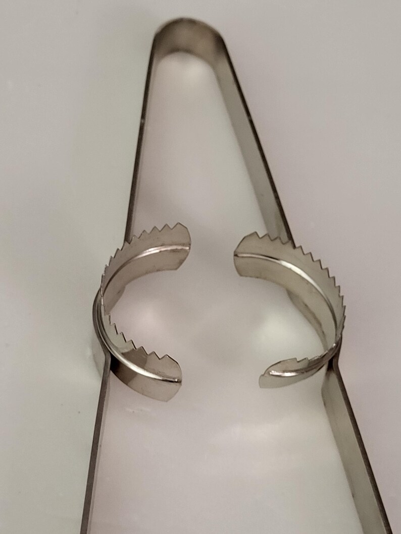 Puede incluir: Un par de pinzas de metal plateado con bordes dentados en los extremos. Las pinzas est&aacute;n abiertas y listas para agarrar algo.
