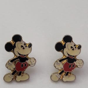 May include: A pair of Mickey Mouse earrings. Each earring features a gold-colored outline with black ears, a cream-colored face and hands, a red shorts, and cream-colored shoes. The earrings are set against a white background.