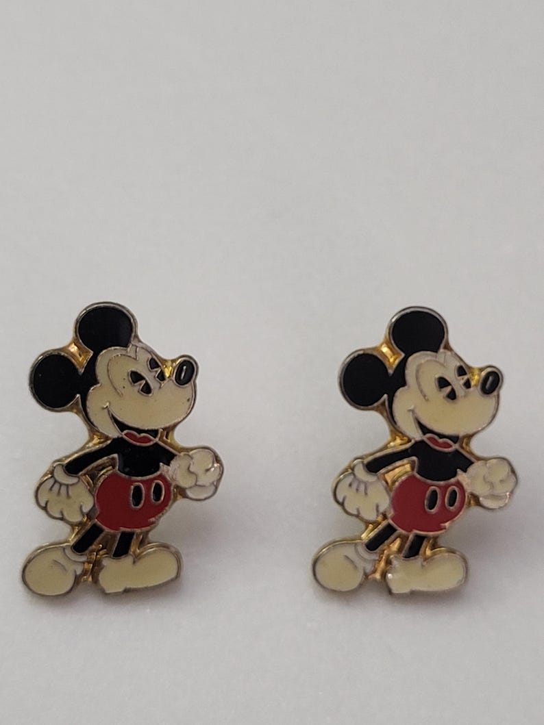 May include: A pair of Mickey Mouse earrings. Each earring features a gold-colored outline with black ears, a cream-colored face and hands, a red shorts, and cream-colored shoes. The earrings are set against a white background.