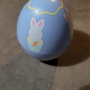 May include: A light blue Easter egg-shaped toy with a yellow wavy design near the top. The egg features a cartoon bunny holding a carrot. The bunny has pink ears and is wearing a light blue outfit.