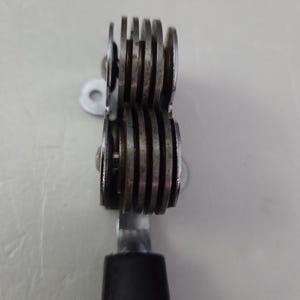May include: A metal tool with two sets of grooved wheels, connected by a metal bar and a black handle. The wheels are made of a silver-colored metal and are stacked closely together. The tool appears to be used for cutting or scoring.