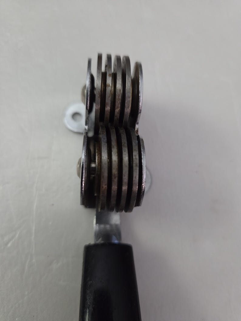 May include: A metal tool with two sets of grooved wheels, connected by a metal bar and a black handle. The wheels are made of a silver-colored metal and are stacked closely together. The tool appears to be used for cutting or scoring.