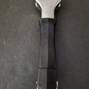 May include: A stainless steel grill scraper with a black handle. The scraper has a wide, flat blade and a sturdy, angled handle for easy grip. A small metal loop is at the end of the handle.