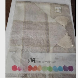 May include: A packaged table runner with a beige and brown checked design. The runner has two beige tassels and is labelled with the text "la. TABLE TOP COORDINATES" along with a row of colourful watercolour splotches.