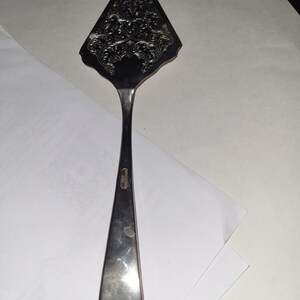May include: A silver cake server with an ornate design. The server has a pointed blade and a long, slender handle.