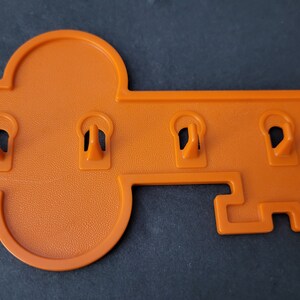 May include: Orange key-shaped wall-mounted key holder with four keyhole-shaped slots. The key holder is made of plastic and has a textured surface. The key holder is designed to be mounted on a wall.