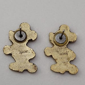 May include: Two gold-colored Mickey Mouse-shaped stud earrings. The backs of the earrings are visible, showing the post and the Disney logo. The earrings have a textured surface.
