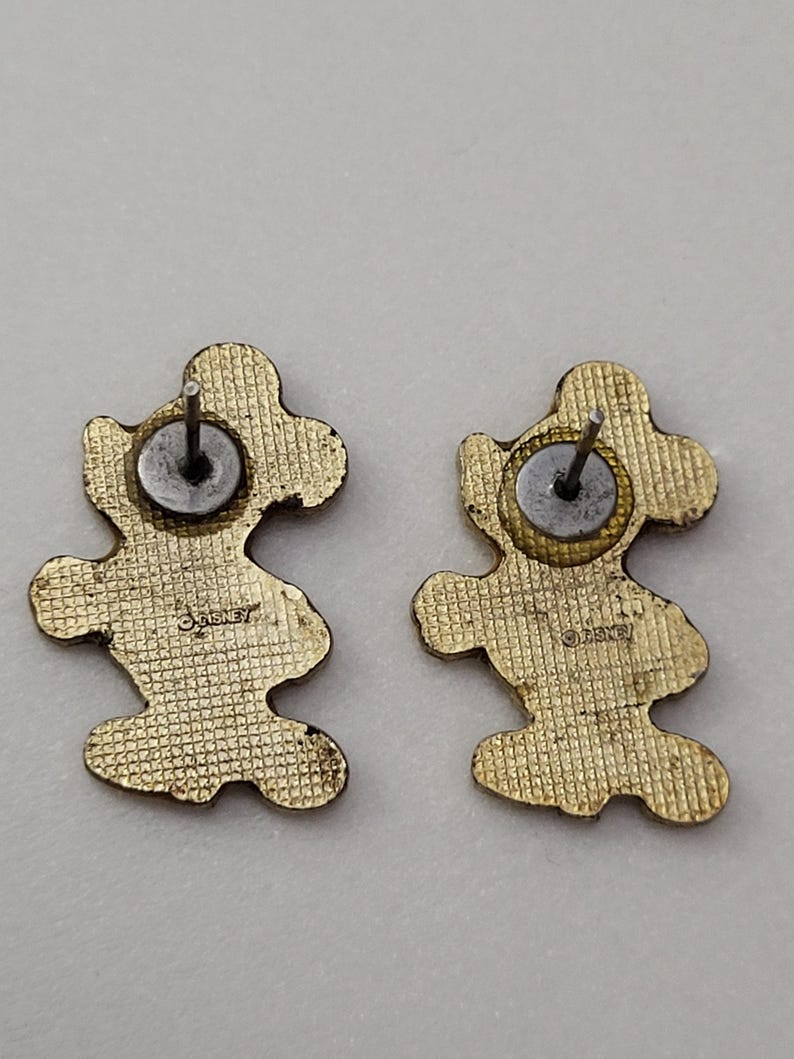 May include: Two gold-colored Mickey Mouse-shaped stud earrings. The backs of the earrings are visible, showing the post and the Disney logo. The earrings have a textured surface.