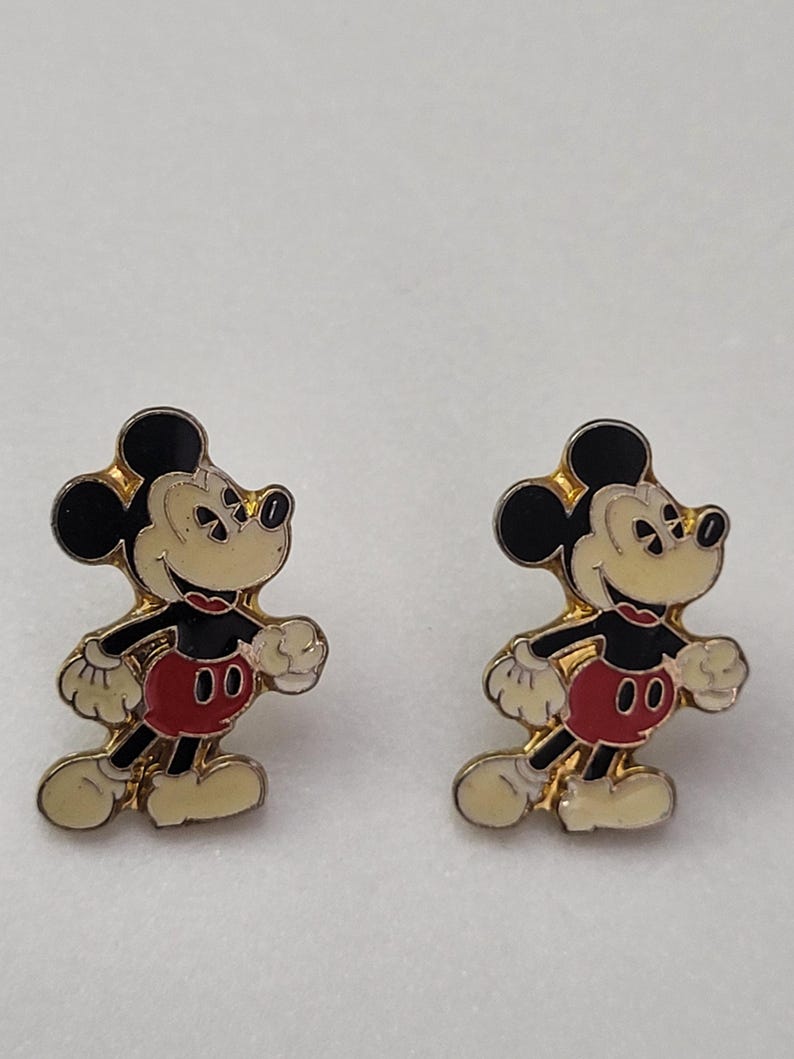 May include: Pair of Mickey Mouse enamel pins. Each pin features Mickey Mouse in a walking pose, with black ears, a cream-colored face, red shorts, and white gloves. The pins have a gold-colored border.