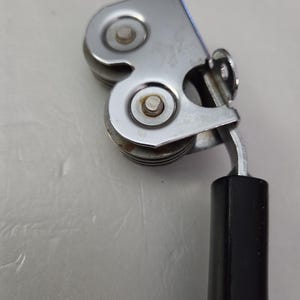 May include: A close-up of a vintage can opener. The can opener is made of shiny, silver-colored metal with two small, round cutting wheels. It has a black, rectangular handle.