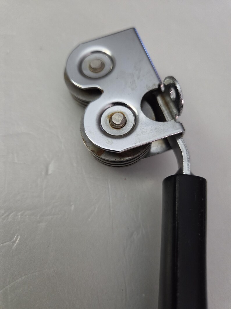 May include: A close-up of a vintage can opener. The can opener is made of shiny, silver-colored metal with two small, round cutting wheels. It has a black, rectangular handle.