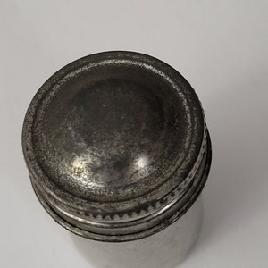 May include: A silver metal container with a screw-on lid. The lid has a raised, rounded top.