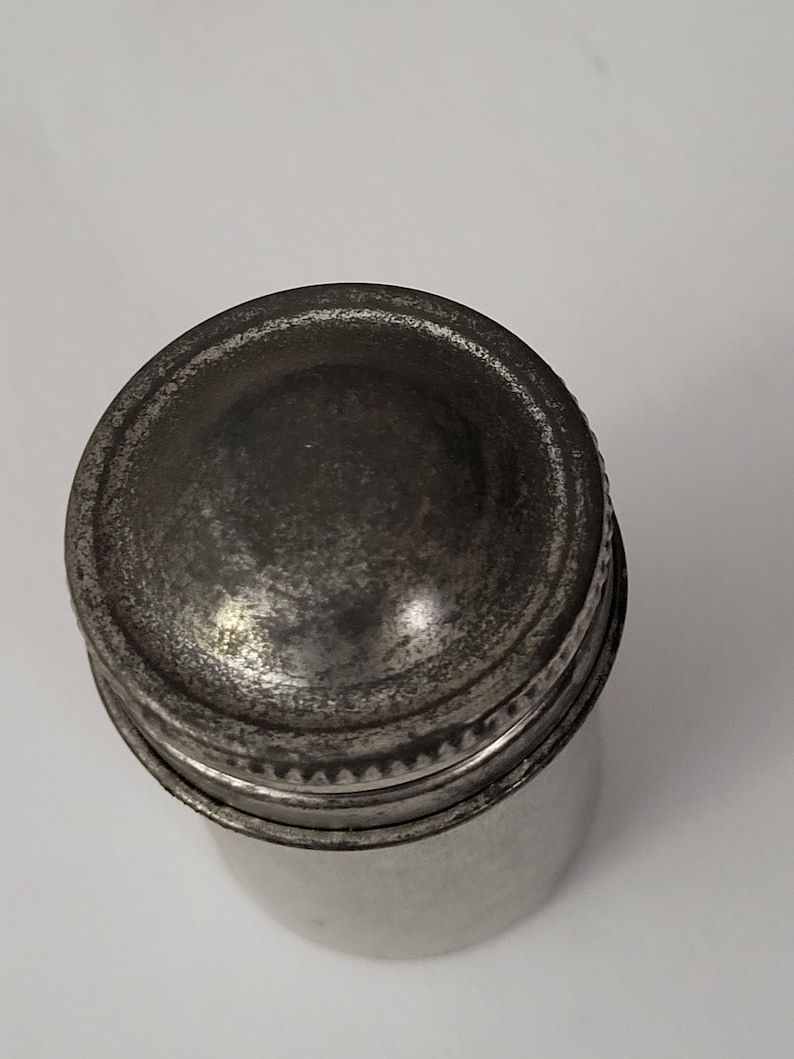 May include: A silver metal container with a screw-on lid. The lid has a raised, rounded top.