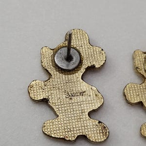 May include: Gold-toned Mickey Mouse stud earrings. The earrings are shaped like Mickey Mouse with a textured surface. The back of one earring shows the Disney logo. The earrings have a post and a metal backing.