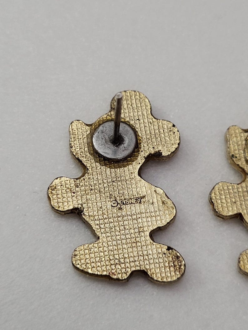 May include: Gold-toned Mickey Mouse stud earrings. The earrings are shaped like Mickey Mouse with a textured surface. The back of one earring shows the Disney logo. The earrings have a post and a metal backing.