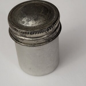 May include: A silver metal container with a screw-on lid. The container is cylindrical and has a slightly domed lid.