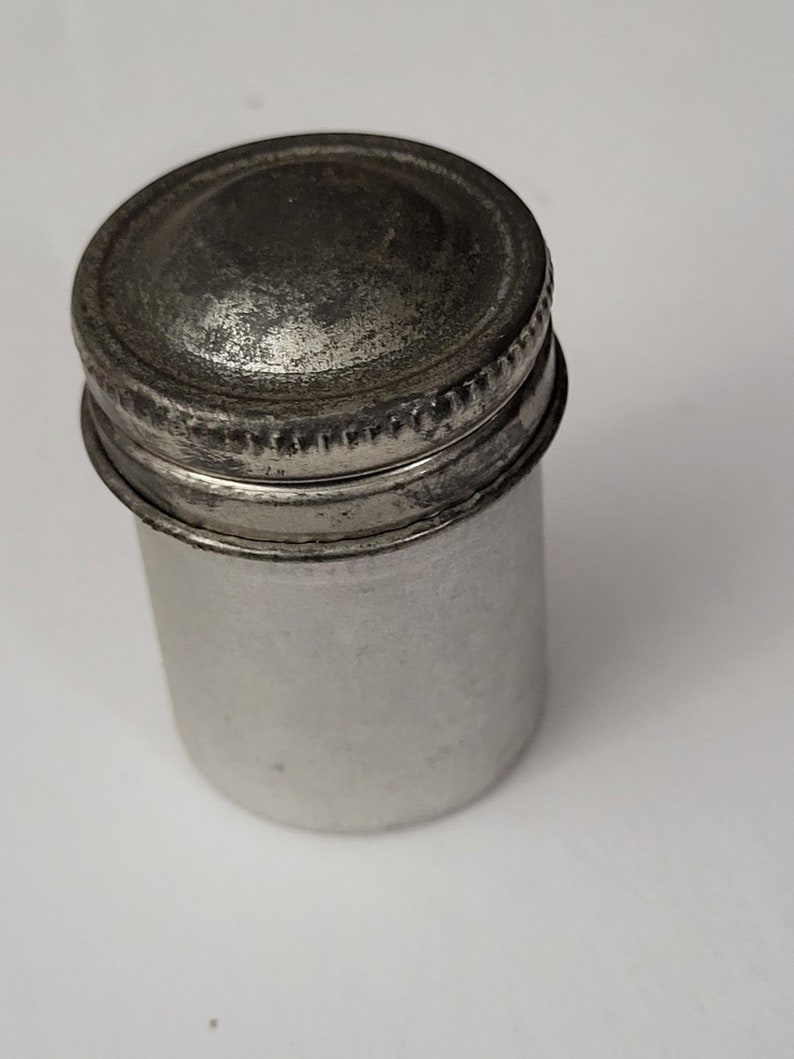 May include: A silver metal container with a screw-on lid. The container is cylindrical and has a slightly domed lid.