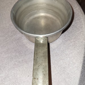 May include: A silver metal measuring cup with a handle. The cup is empty and has a smooth, shiny surface.