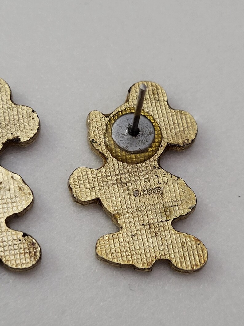 May include: Gold-toned Mickey Mouse stud earrings. The earrings are in the shape of Mickey Mouse with a textured surface. The back of the earrings have a post and a circular backing. The word "Disney" is stamped on the back of the earrings.