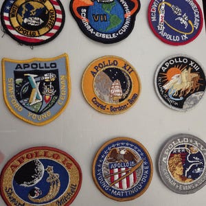May include: A collection of nine embroidered NASA Apollo mission patches arranged in a grid. The patches feature circular and shield shapes with detailed space-themed illustrations, mission names, and astronaut surnames stitched in various colors.