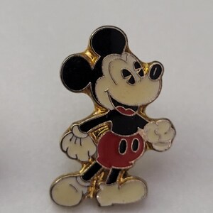 May include: A vintage Mickey Mouse enamel pin. The pin features Mickey Mouse in his classic pose, with black ears, a black body, red shorts, and white gloves and shoes. The pin has a gold-tone border.