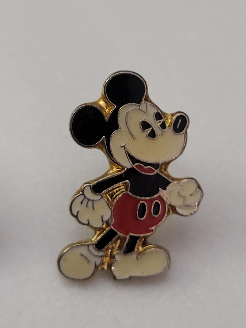 May include: A vintage Mickey Mouse enamel pin. The pin features Mickey Mouse in his classic pose, with black ears, a black body, red shorts, and white gloves and shoes. The pin has a gold-tone border.