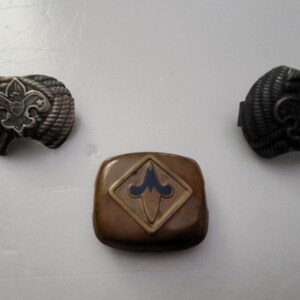 May include: Three vintage Boy Scout insignia pins. Two are silver-toned, with a fleur-de-lis emblem on a textured background. The third is a brown square with a blue and gold fleur-de-lis design.