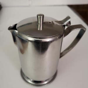 May include: A silver stainless steel creamer with a hinged lid and a handle. The creamer has a decorative band around the bottom.