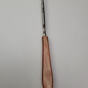 May include: A vintage crochet hook with a metal shaft and a curved hook at the top. The handle is a light pink, elongated shape. The hook is designed for crafting and textile work.