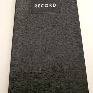 May include: A black record book with a textured cover. The word "RECORD" is printed in silver on a rectangular label at the top. The book has a rectangular shape and appears to be closed.