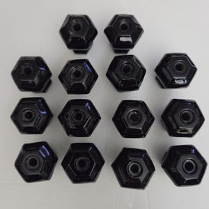 May include: A collection of fourteen black cabinet knobs arranged in four rows. Each knob features a glossy, hexagonal shape with a circular center hole for mounting. The knobs are uniform in size and color, presenting a smooth, reflective surface.