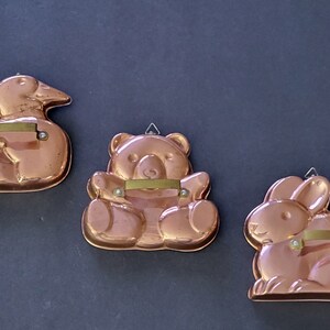 May include: Three vintage copper cake molds in the shapes of a duck, a bear, and a rabbit. Each mold has a brass handle.