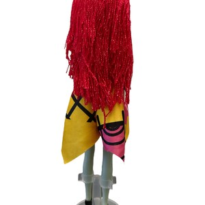 K&ouml;nnte beinhalten: Eine Puppe mit langen, roten Garnhaaren, die ein gelbes und pinkfarbenes Kleid tr&auml;gt. Die Puppe steht auf einem durchsichtigen Plastikst&auml;nder.
