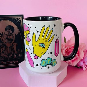 May include: White ceramic mug with a black handle and interior, featuring colorful illustrations of a yellow hand with an eye, mushrooms, and crystals. A tarot card with the text "The Empress" is on the left. The mug is on a pink hexagonal stand.